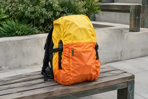 Housse de sac à dos jaune et orange installée sur un sac urbain posé sur un banc en bois